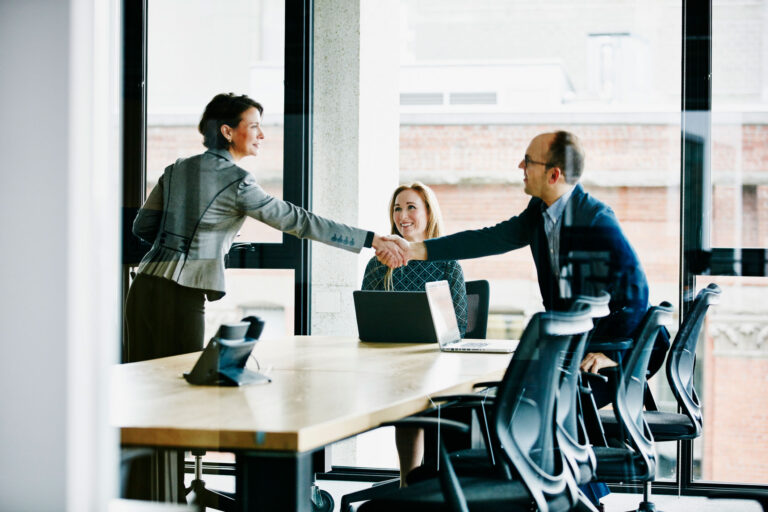 professional woman and man shaking hands across a conference room table with a second woman smiling at them